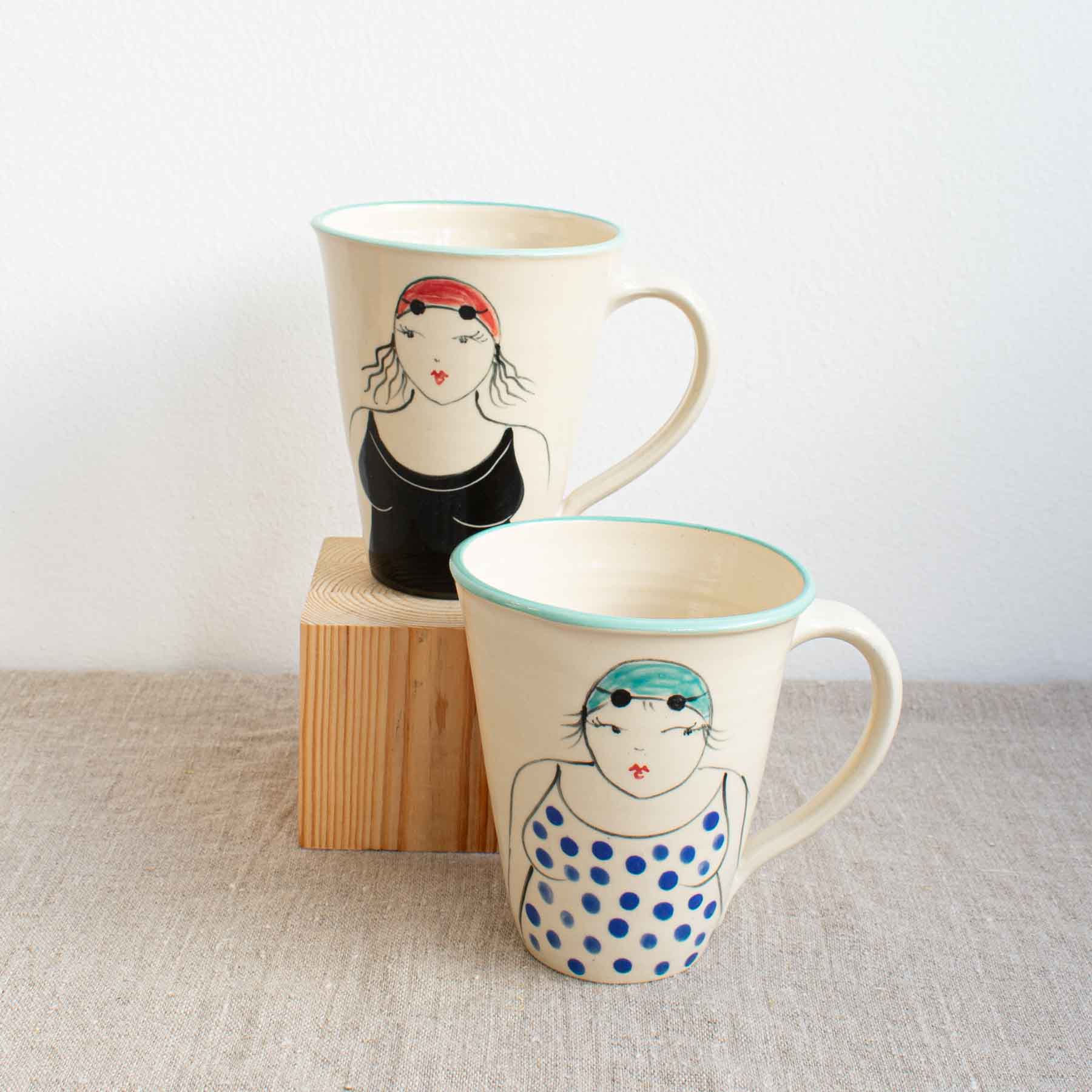 Two handcrafted cream large ceramic mugs with a blue rim and painted with a lady in swimming costume, hat and goggles by Lucie Sivicka, sitting on a linen table cloth