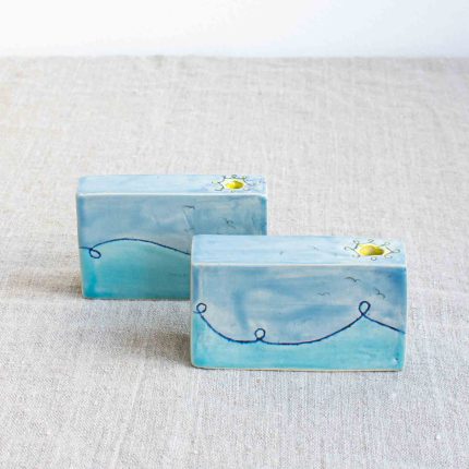 air of rectangular block stem vases decorated with a blue wave design and yellow sun around the opening, handcrafted by Michelle Foote, sitting on a linen table cloth