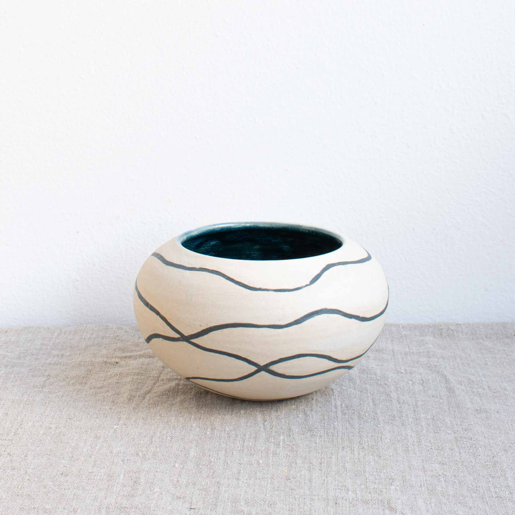 Ceramic pebble pot with matt white outside decorated with deep blue wavy lines and deep blue glazed inner, handcrafted by Michelle Foote, sitting on a linen table cloth