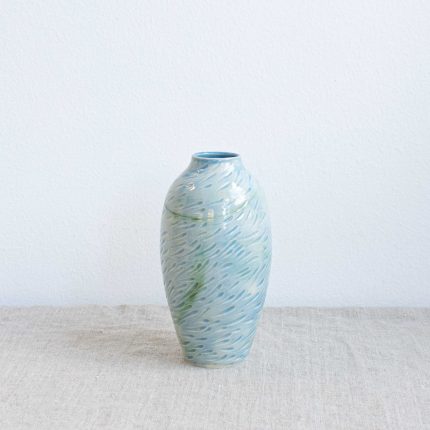 Tall slim ceramic vase with carved design resembling a shoal of fish and finished with a high shine pale sea blue glaze, handcrafted by Michelle Foote, sitting on a linen table cloth