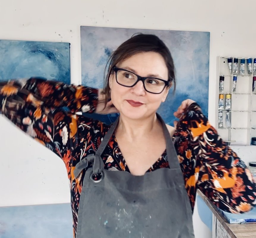 Woman in glasses with floral shirt and apron in studio with paintings on white wall