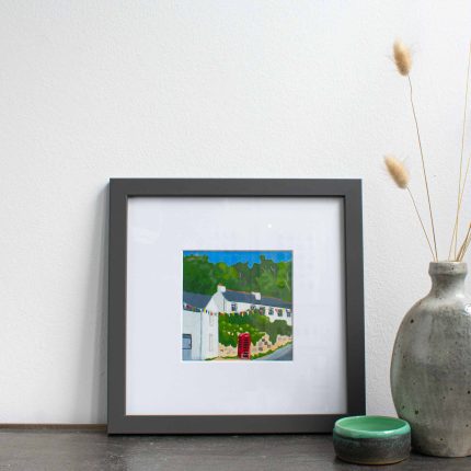 Painting of cottages and red phone box with blue sky in white mount and grey frame on table with vase by Karen Hemsley-Biggs