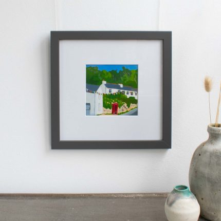 Painting of cottages and red phone box with blue sky in white mount and grey frame on wall over table with vase by Karen Hemsley-Biggs