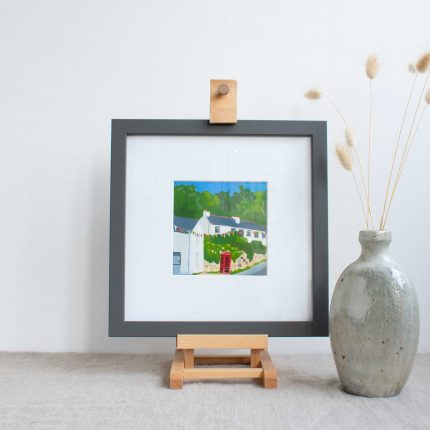 Painting of cottages and red phone box with blue sky in white mount and grey frame, on easel on linen table cloth with vase by Karen Hemsley-Biggs