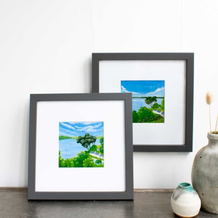 Pair of paintings of trees and cottages on the coast with blue sky in white mount and grey frame on wall over table with vase by Karen Hemsley-Biggs