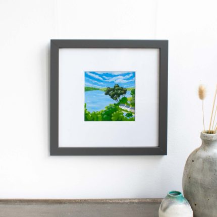 Painting of trees and cottages on the coast with blue sky in white mount and grey frame on wall over table with vase by Karen Hemsley-Biggs