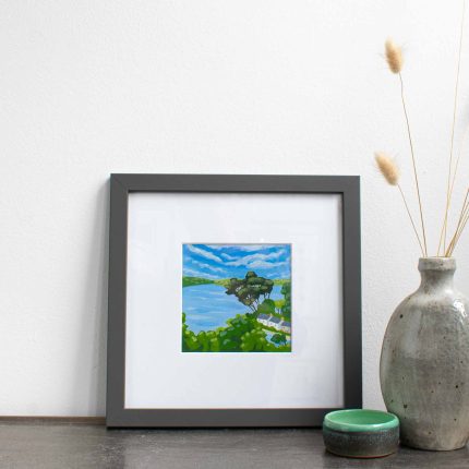 Painting of trees and cottages on the coast with blue sky in white mount and grey frame on table with vase by Karen Hemsley-Biggs