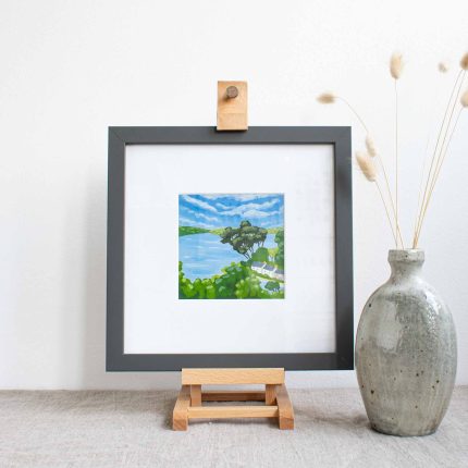 Painting of trees and cottages on the coast with blue sky in white mount and grey frame, on easel on linen table cloth with vase by Karen Hemsley-Biggs