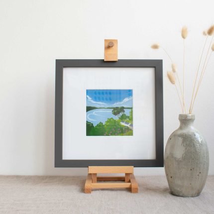 Painting of trees and cottages on the coast with blue sky in white mount and grey frame on easel on linen table cloth table with vase by Karen Hemsley-Biggs