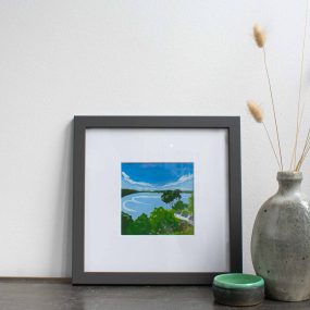 Painting Of Trees And Cottages On The Coast With Blue Sky In White Mount And Grey Frame On Table With Vase By Karen Hemsley-Biggs