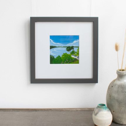 Painting of trees and cottages on the coast with blue sky in white mount and grey frame on wall over table with vase by Karen Hemsley-Biggs