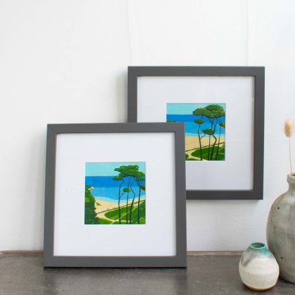 Pair of paintings of trees on the coast with blue sky in white mount and grey frame on wall over table with vase by Karen Hemsley-Biggs