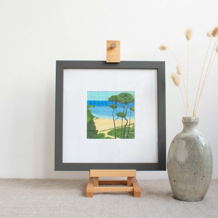 Painting of trees on the coast with blue sky in white mount and grey frame, on easel on linen table cloth with vase by Karen Hemsley-Biggs