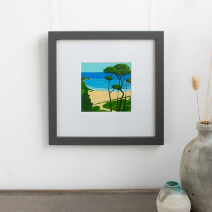 Painting of trees on the coast with blue sky in white mount and grey frame on wall over table with vase by Karen Hemsley-Biggs