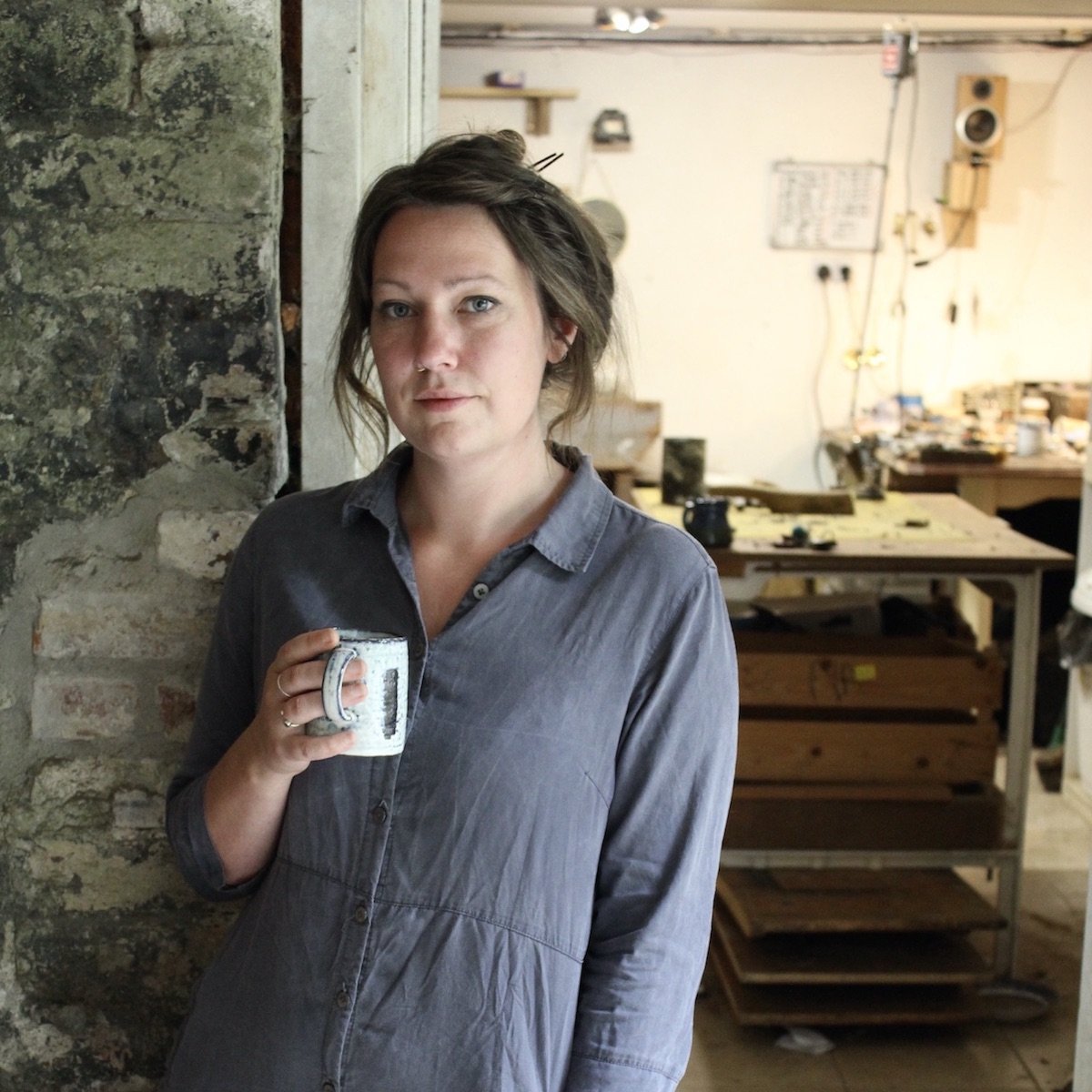 Woman holding a coffee cup against a stone wall with workshop in background