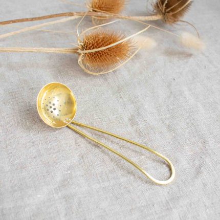 Long handled brass olive scoop handcrafted by Razmaker on a linen tablecloth with teasels